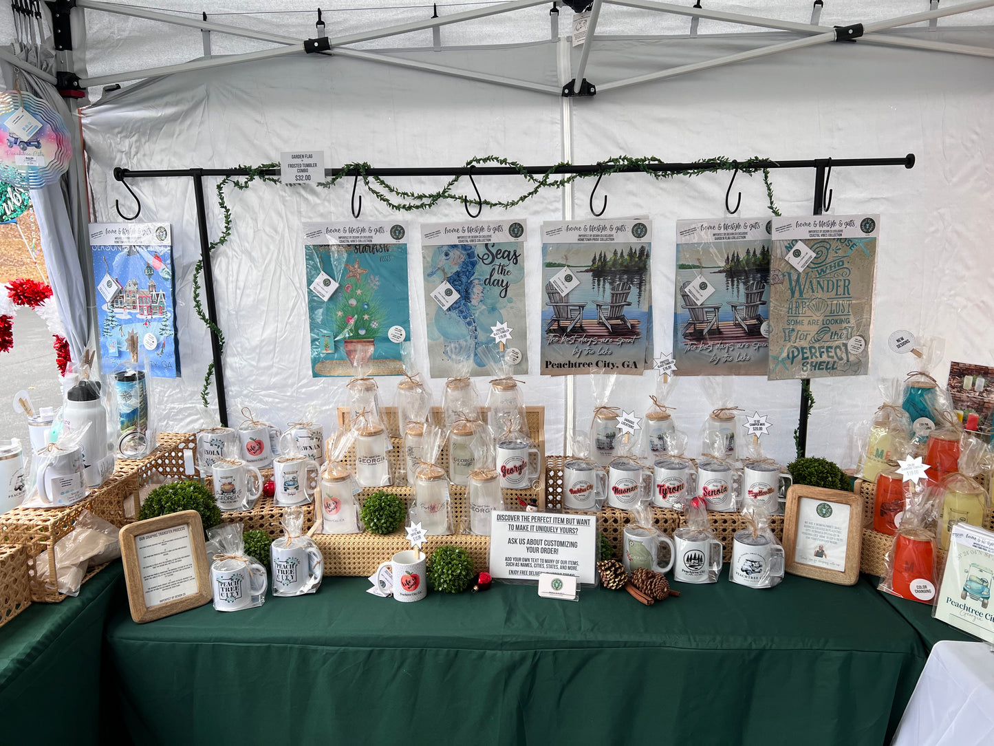 Vendors' table with various products under a white tent, featuring mugs and decorative items and personalized garden flags.  | imperfect by design co