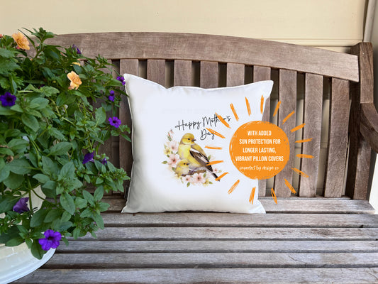 White decorative pillow advertising pillow cover with a female Goldfinch and it's nestling sitting on a branch with pink flowers. Text on pillow reads "Happy Mother's Day' . Placed on a porch bench flanked by a trailing petunia in a white pot. Sun shaped blurb reads 'with added sun protection for longer lasting vibrant pillow covers' | imperfect by design co