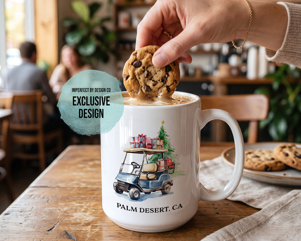 A white ceramic mug with a colorful illustration of a golf cart carrying a Christmas tree and presents, labeled 'PALM DESERT, CA'. A hand is placing a chocolate chip cookie into a cup of coffee inside a cozy cafe setting.