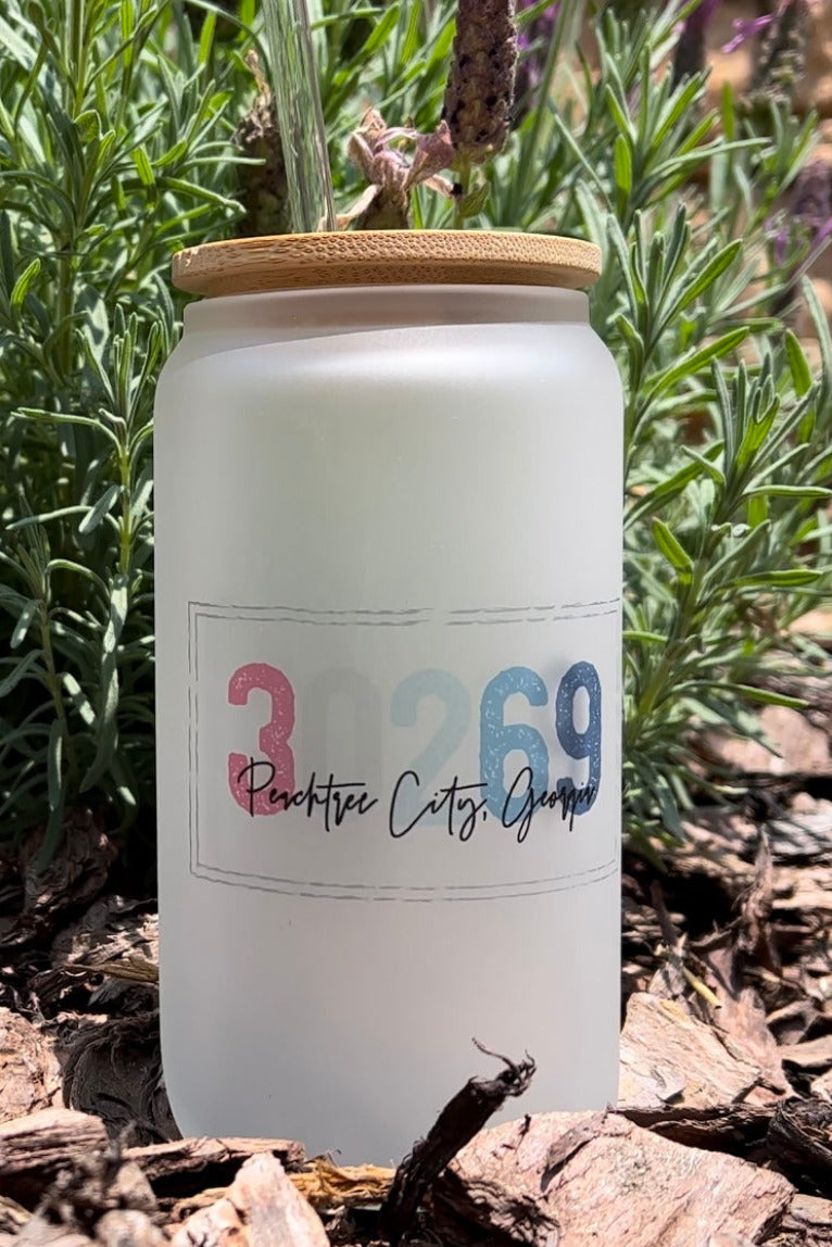 Frosted glass tumblers with bamboo lid with clear straw, sitting outdoors on mulch with green plants behind it. Tumbler has a red, white and blue text on right tumbler reads '30269 Peachtree City, Georgia' . imperfect by design co by design co exclusive