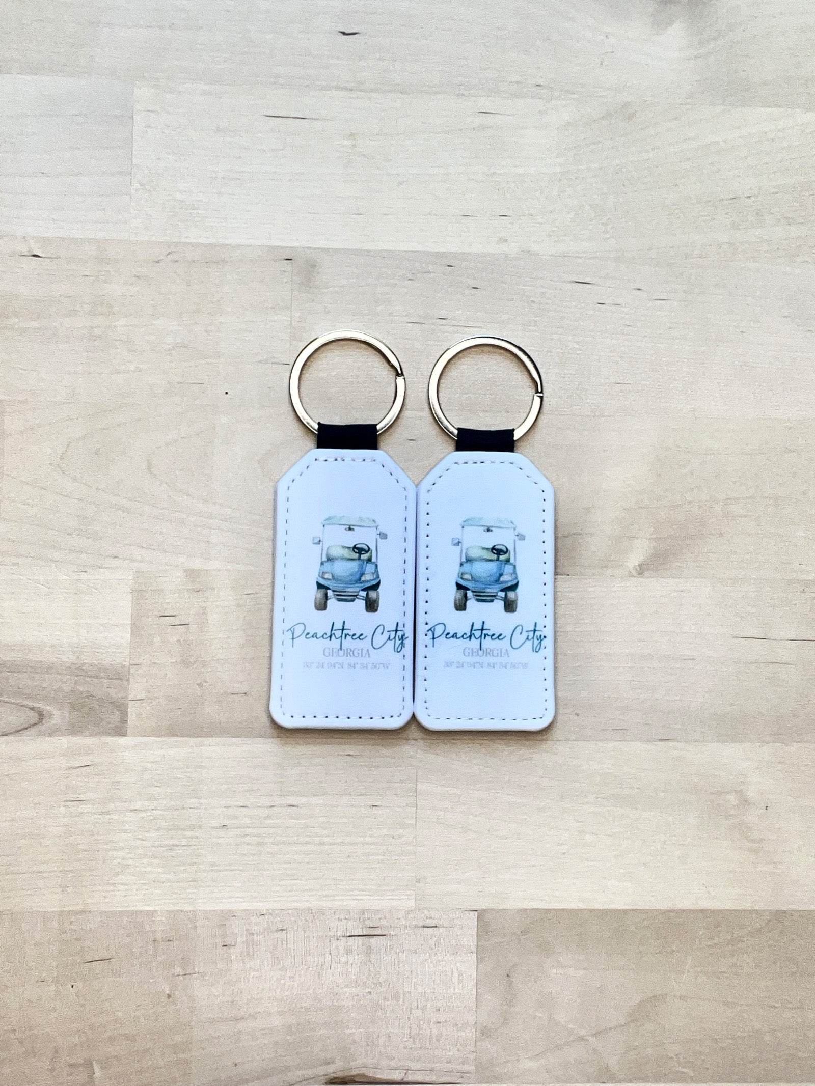 Exclusive design on 2 white faux leather keychain, with a blue golf cart and green text, including coordinates, and the text 'Peachtree City, GA'.  View of both sides on a wood background| imperfect by design co 