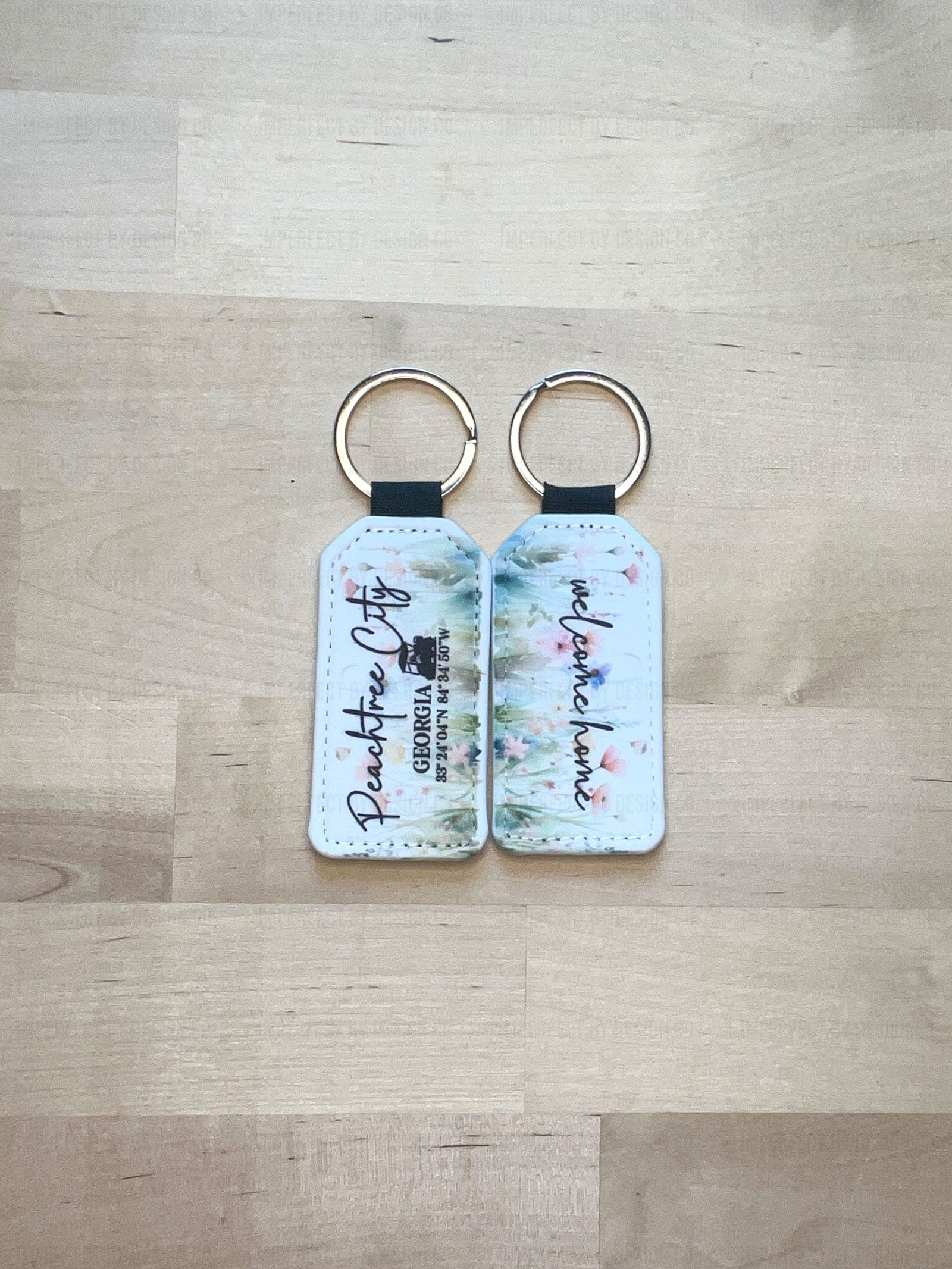 Two Exclusive Peachtree City Wildflower and Poppies Keychain view of front and back, Text on Front features a golf cart and reads "Peachtree City Georgia with Peachtree City coordinates" Back of Key Chain reads "welcome home" on a pale pink background  | imperfect by design co 