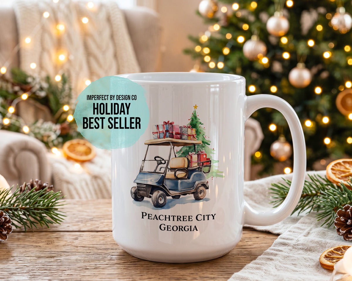 A white ceramic mug with a custom golf cart and Christmas tree illustration, featuring the text 'PEACHTREE CITY GEORGIA'. The mug is displayed on a wooden table with a festive holiday background containing a decorated Christmas tree, pine branches, and warm string lights."Holiday Bestseller" on blue text blurb.