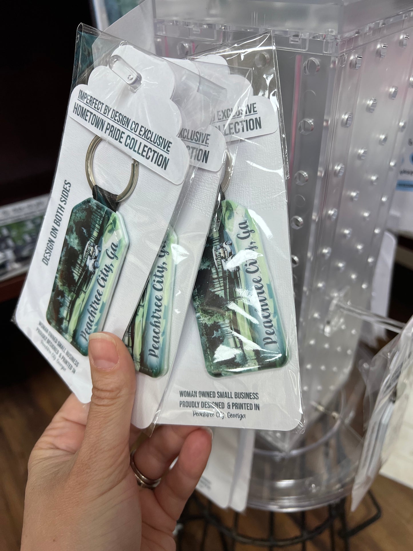 A faux leather key chain featuring a printed design with a golf cart scene, with the text 'Peachtree City GA' on a key ring. Placed a retail stand being and being held up by a hand | imperfect by design co exclusive design