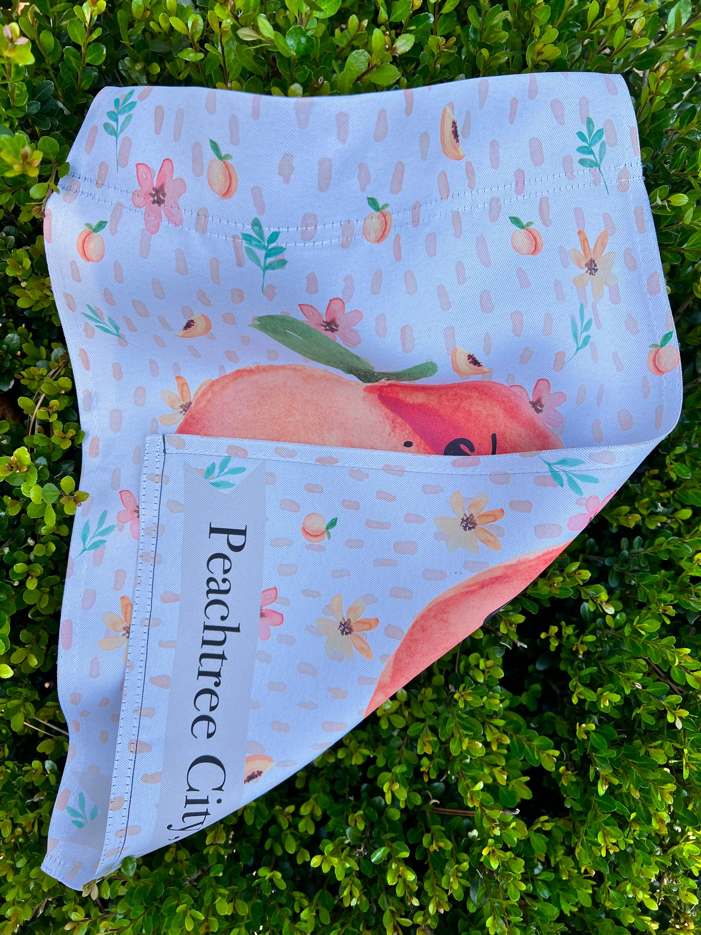 Exclusive imperfect by design co design Personalized decorative garden flag with peach design and text on a white background filled with peaches, peach blooms and leaves. Garden flag is placed on a boxwood background. Text on Flag reads 'Life is sweet' and 'Peachtree City, GA' in black text. The flag is folded in half to display that is printed on both the front and the reverse.