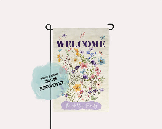 A vertical garden flag displayed on a black metal stand against a pale pink background. The flag has a cream base with a watercolor-style floral bouquet in purple, blue, yellow, and pink, and the word 'WELCOME' in dark purple at the top.
