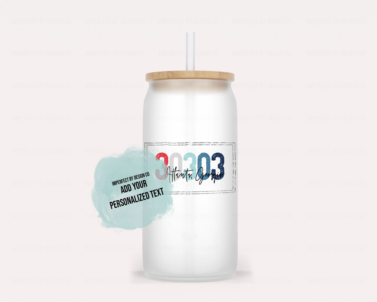 Personalized frosted glass tumbler with a bamboo lid and clear straw, shown on a pale pink background. The text on the tumbler reads '30303' in red and blue and the words 'Atlanta Georgia' in script, plus a light blue text bubble that says 'imperfect by design co add your personalized text'