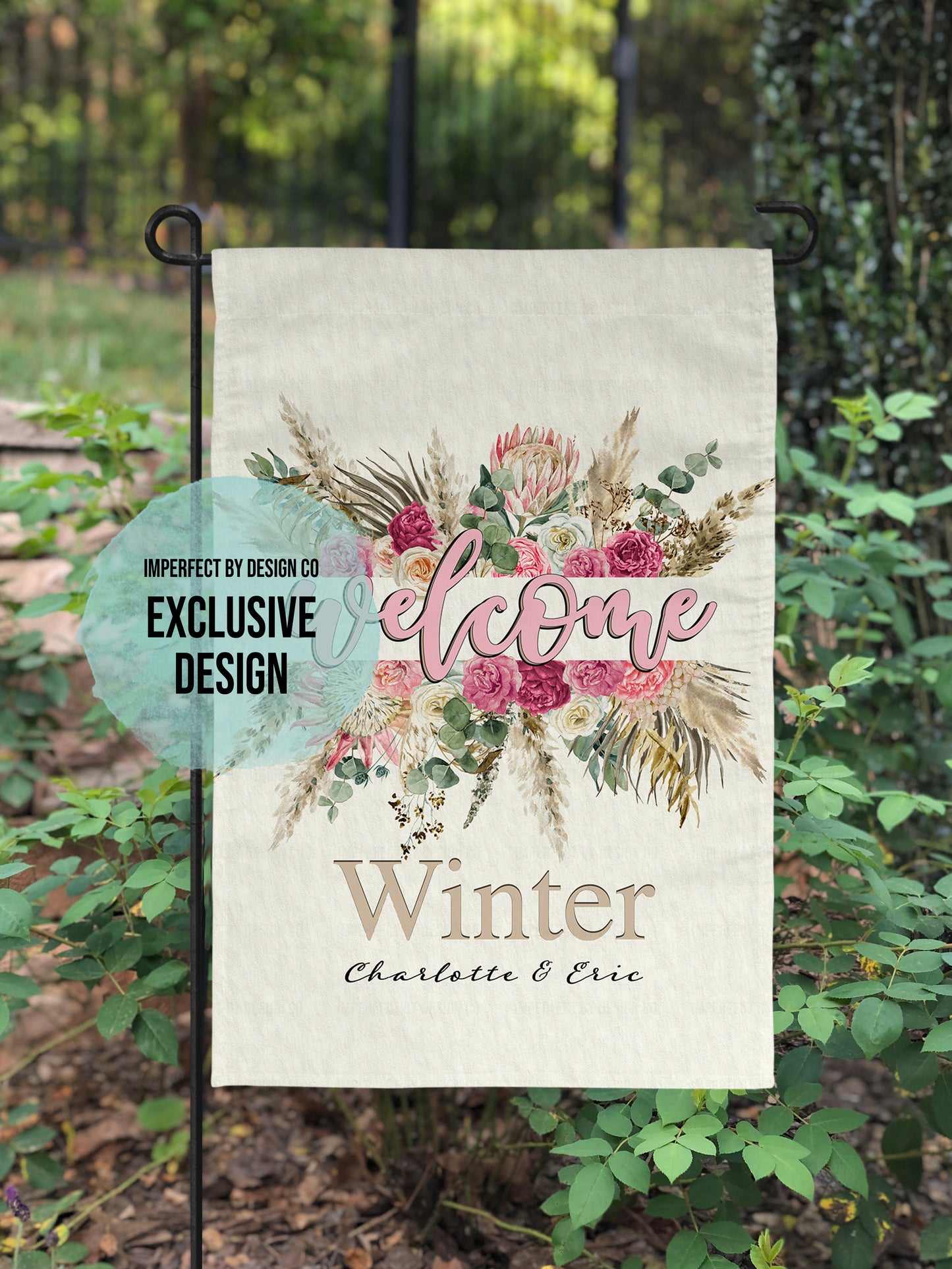 imperfect by design co exclusive 'Welcome' garden flag featuring split floral design and personalization. Placed on garden flag stand in lush garden setting.
