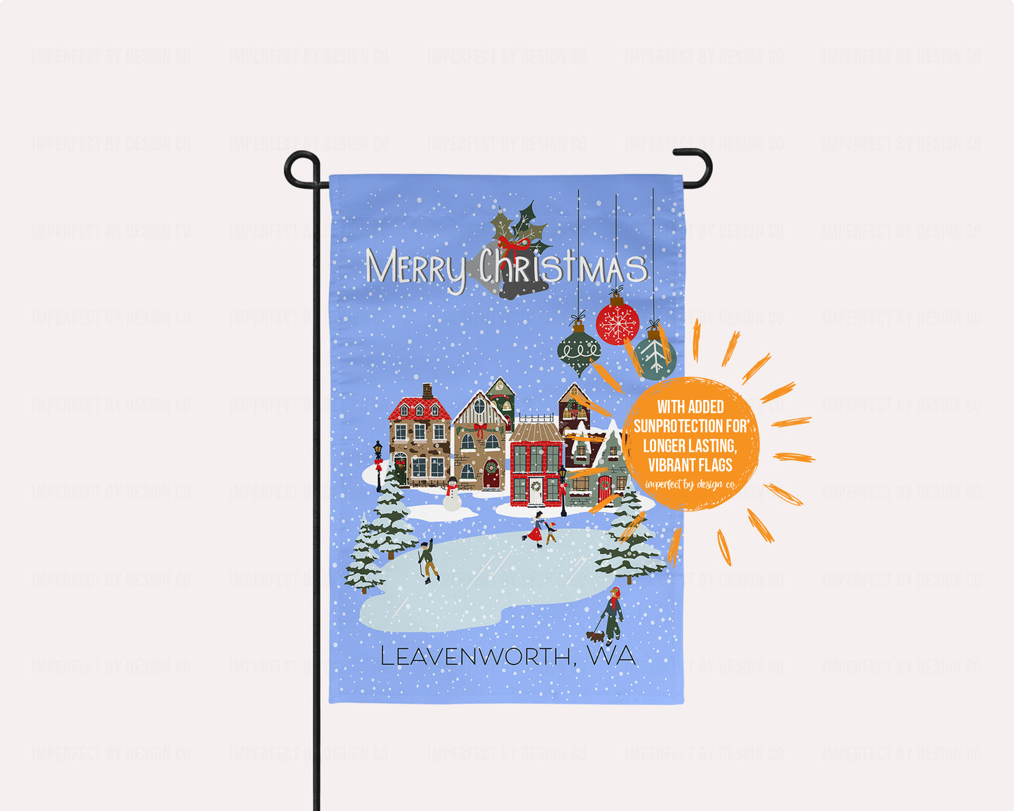 Christmas-themed garden flag with a snowy village scene and 'Merry Christmas' text, featuring Leavenworth, WA. Sun shaped blurb on flag reads with added protection for longer lasting vibrant flags. Placed on a pale pink background. | imperfect by design co exclusive design. The flag is placed flag stand, on a pale pink background. 