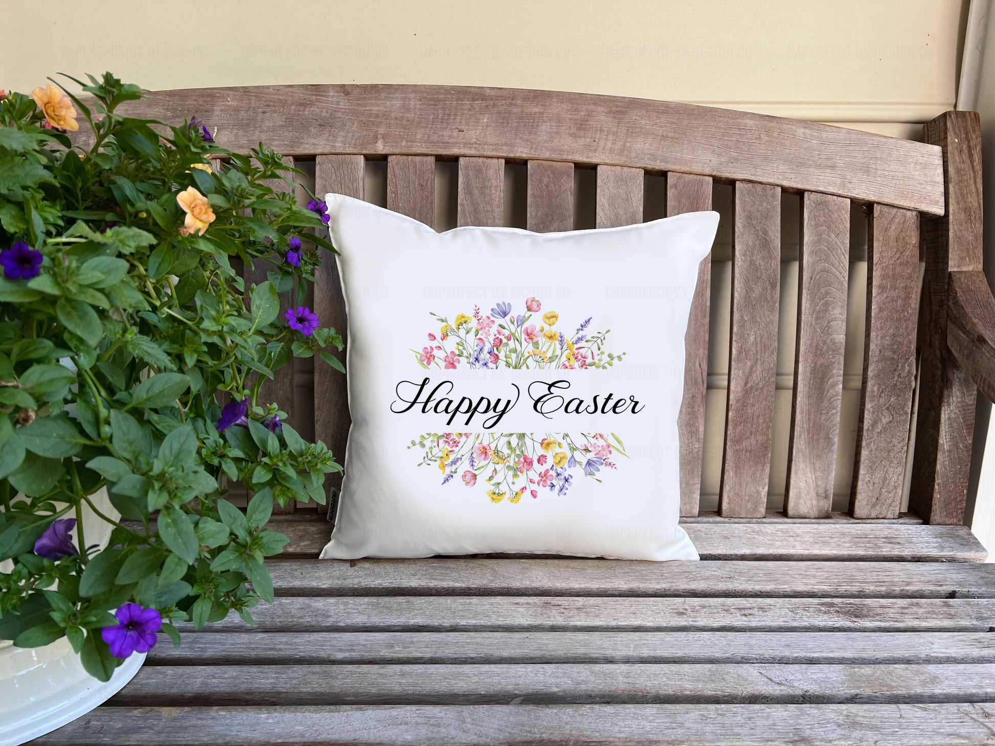 White decorative pillow advertising pillow cover with 'Happy Easter' text on a wooden bench with flowers.