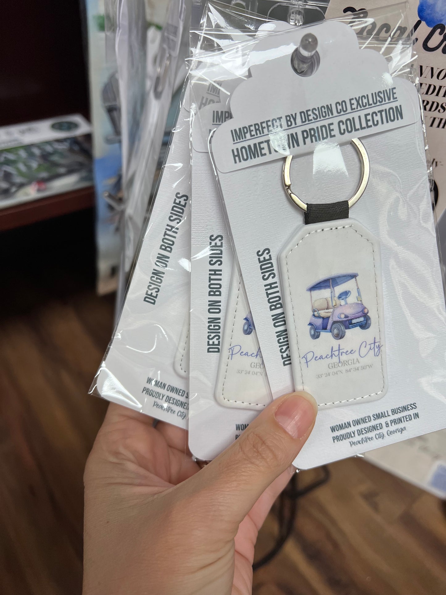 Several retail packaged white faux leather keychain with a blue golf cart and purple  text, including coordinates, and the text 'Peachtree City, GA'. On a retail stand held up by a hand | imperfect by design co exclusive design 
