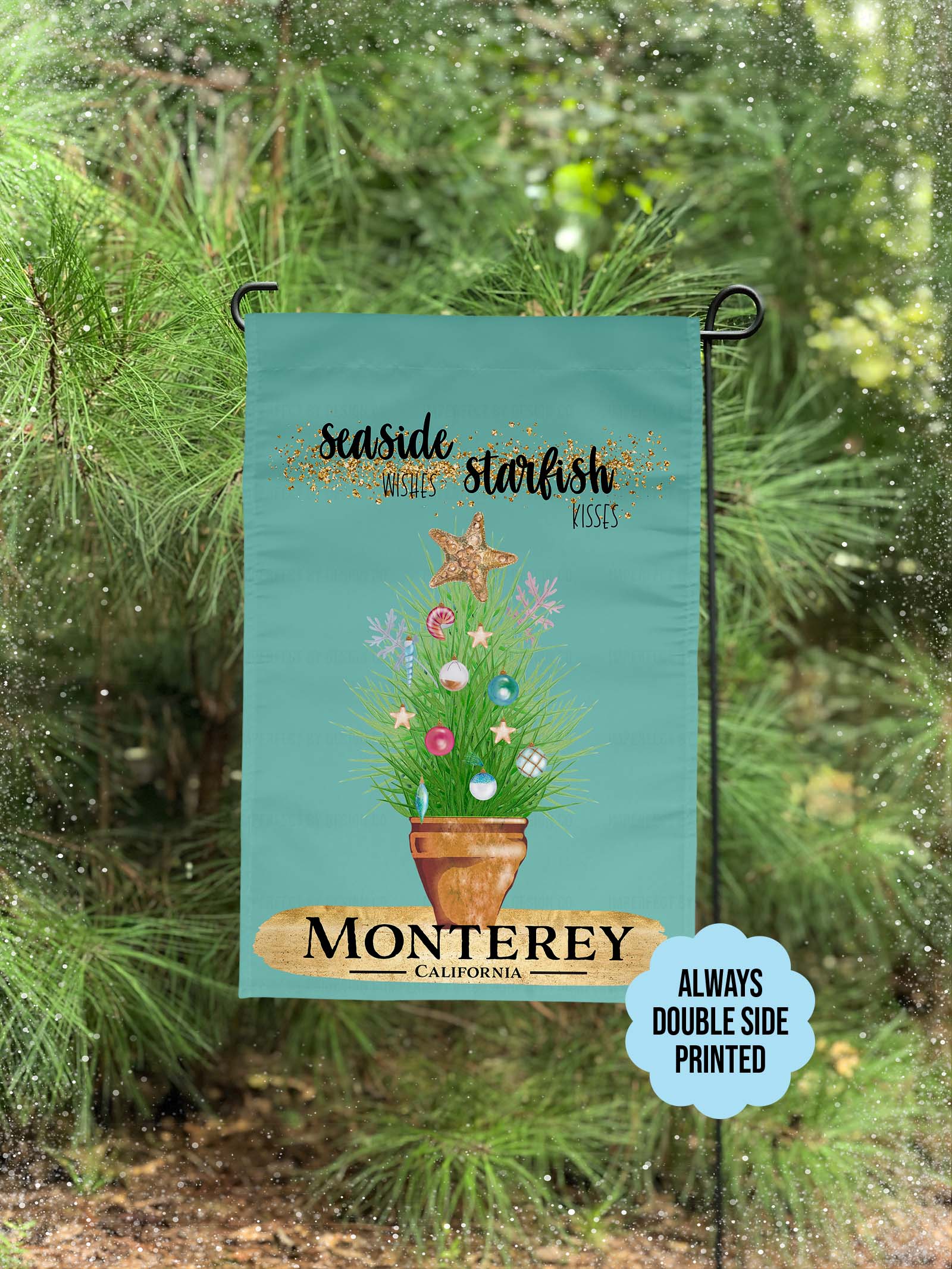 The blue green colored garden flag featuring potted ornamental grass adorned with Christmas tree bulb and topped with a golden starfish. The Flag reads 'Seaside Wishes Starfish Kisses'. Large golden brushstroke below the plant with black bold text that reads 'Monterey' 'California'. The flag is on a flag pole. A holiday decorate porch is visible in the background.  A blue scallop shape blurb on the flag reads 'Always double side printed.' A pine tree and snowflakes are visible. 