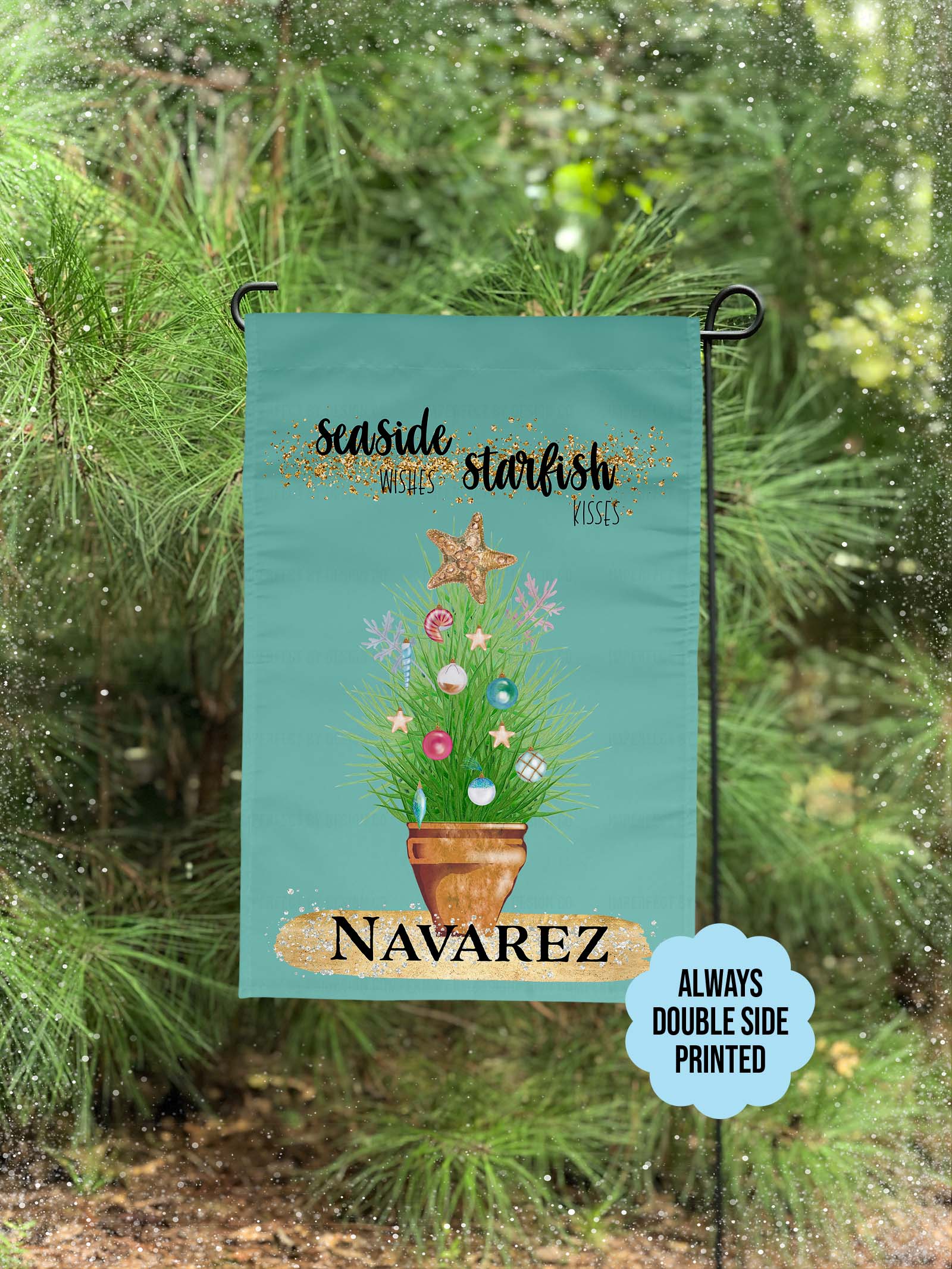 Personalized with a Last Name Flag. The blue green colored garden flag featuring potted ornamental grass adorned with Christmas tree bulb and topped with a golden starfish. The Flag reads 'Seaside Wishes Starfish Kisses'. Large golden brushstroke below the plant with black bold text that reads 'Navarez'. The flag is on a flag pole. A holiday decorate porch is visible in the background.  A blue scallop shape blurb on the flag reads 'Always double side printed.' A pine tree and snowflakes are visible. 