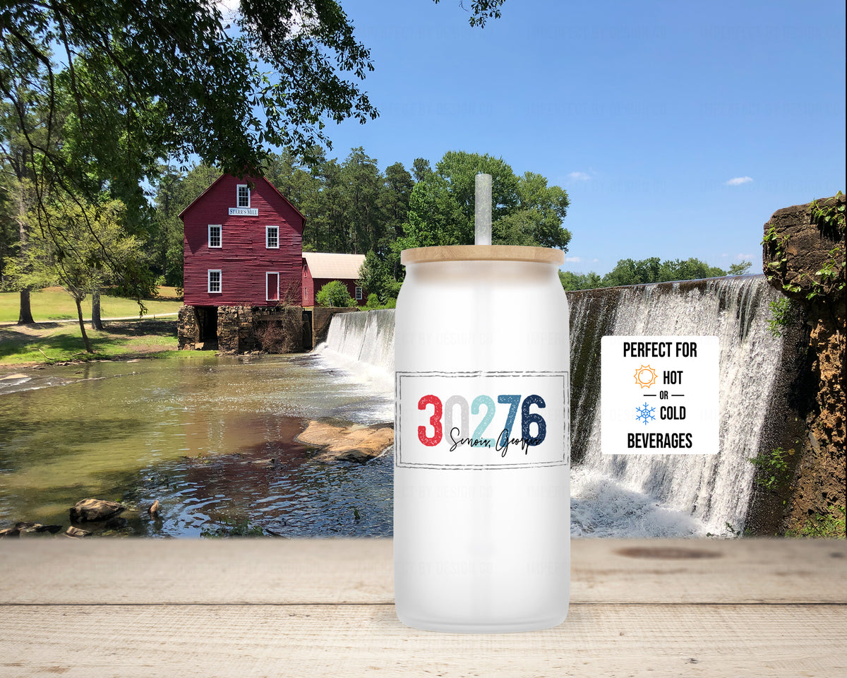 A frosted glass tumbler with a bamboo lid and straw is shown in front in front of Starrs Mill. Text on the tumbler reads '30276 Senoia, Georgia' in red, white and blue and a small text blurb reads 'PERFECT FOR HOT COLD BEVERAGES'.