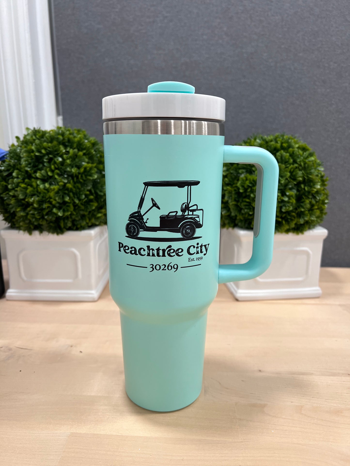 imperfect by design co Exclusive 40oz Turquoise Stainless Steel Tumbler tumbler with black golf cart and text placed on butcher block surface two boxwood topiaries in white planters are visible in the background as well as a gray backdrop.