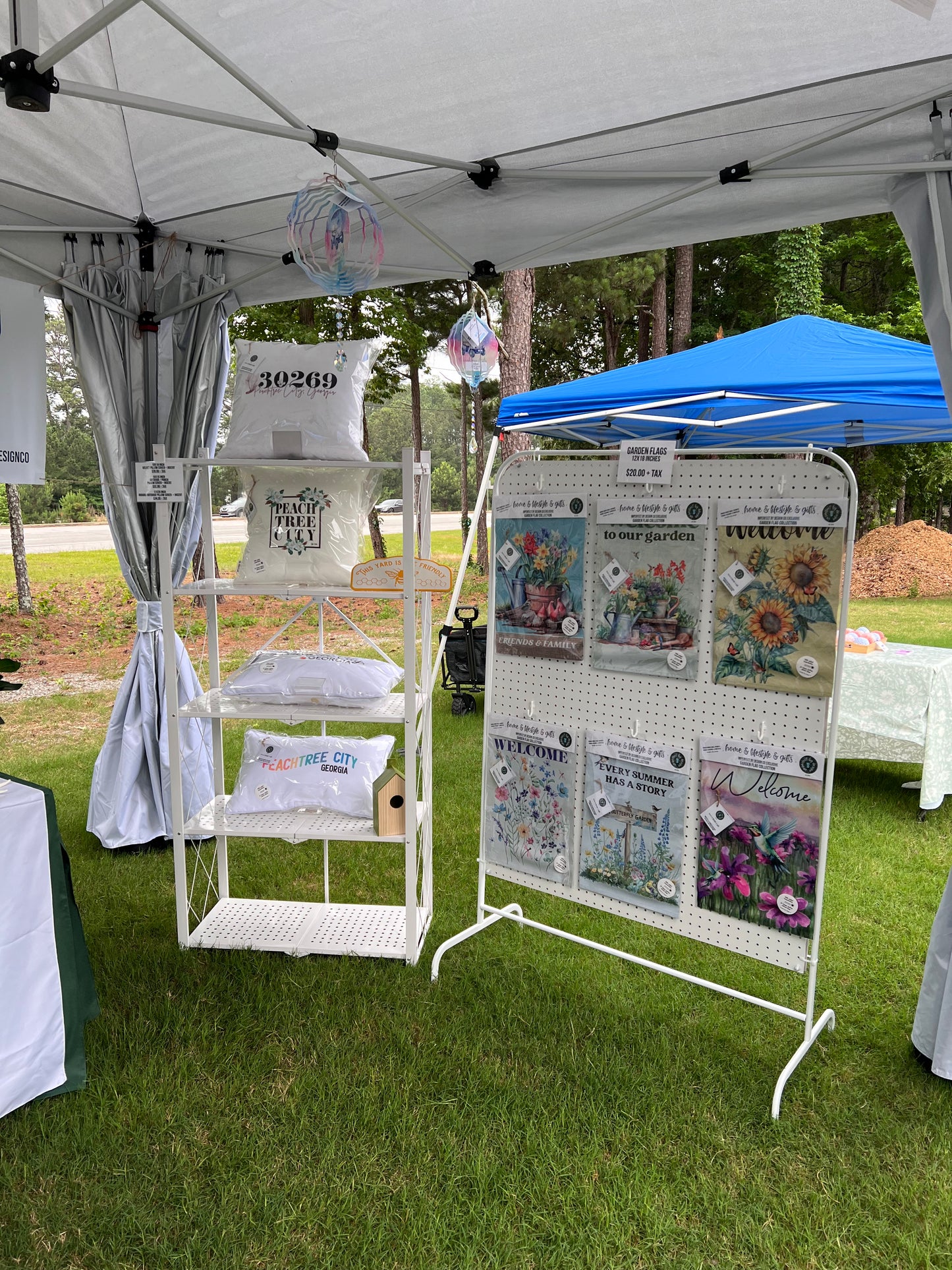 Outdoor Vendor Market setting with imperfect by design co Pillow Cases on a white display stand holding as well as our Garden Flags on a nearby display