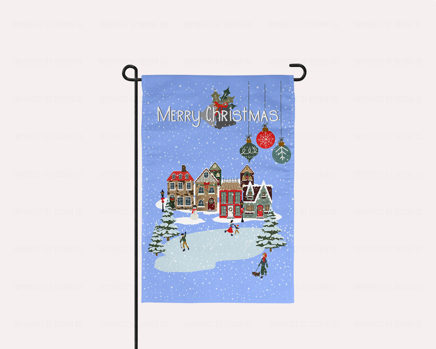 imperfect by design co exclusive design. Christmas-themed garden flag with a winter village scene and 'Merry Christmas' text on a blue background with snowflakes and Christmas tree bulbs. The flag is on a flag stand placed on a pale pink background. This flag is not personalized.