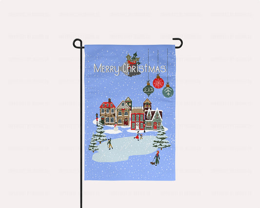 imperfect by design co exclusive design. Christmas-themed garden flag with a winter village scene and 'Merry Christmas' text on a blue background with snowflakes and Christmas tree bulbs. The flag is on a flag stand placed on a pale pink background. This flag is not personalized.