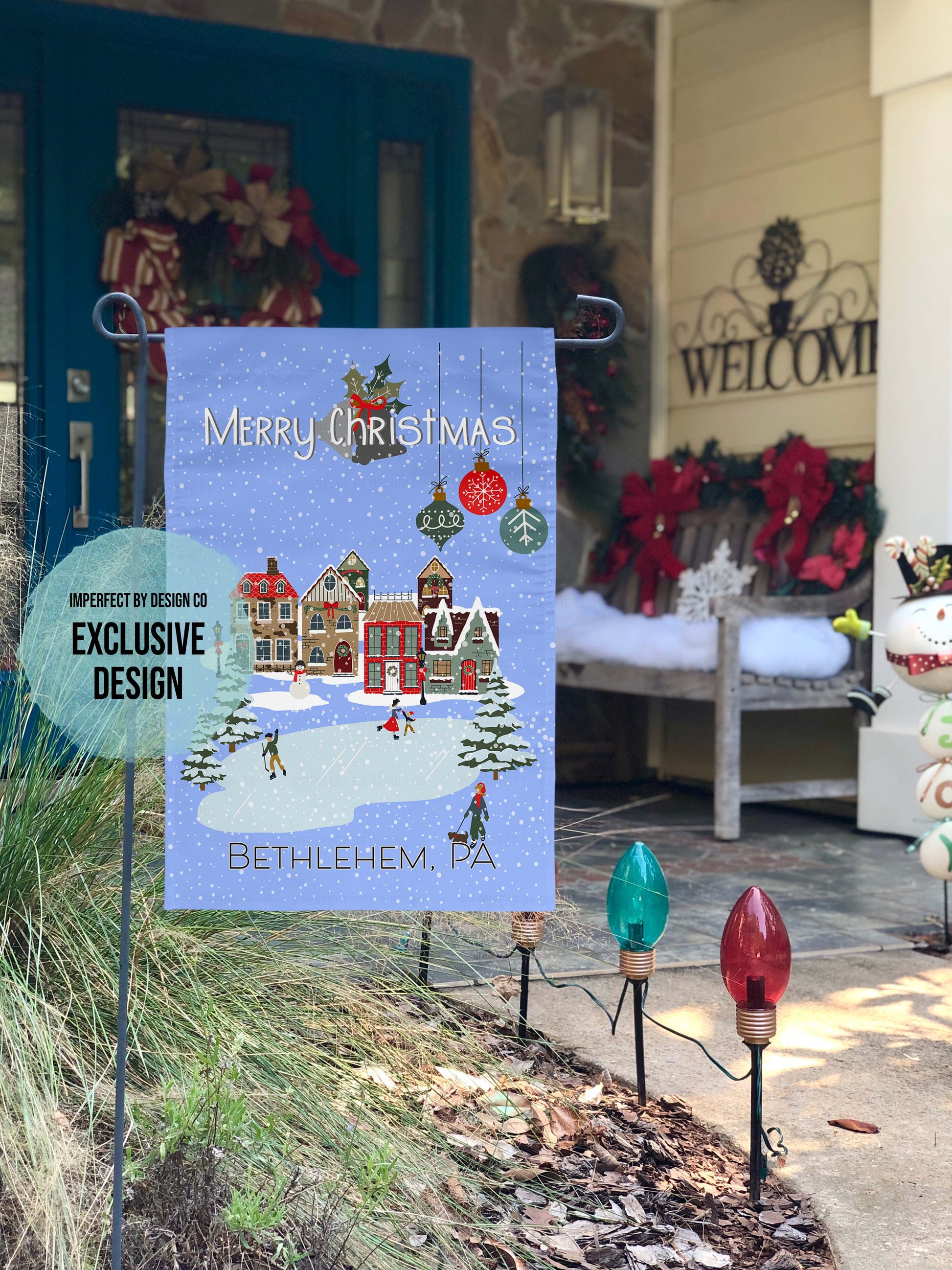 A Christmas-themed garden flag with a snowy village scene and text that reads 'Merry Christmas' personalized with the text, featuring Bethlehem, PA.  The teal blurb on flag reads  imperfect by design co exclusive design. Christmas-themed garden flag with a winter village design in front of a decorated porch.