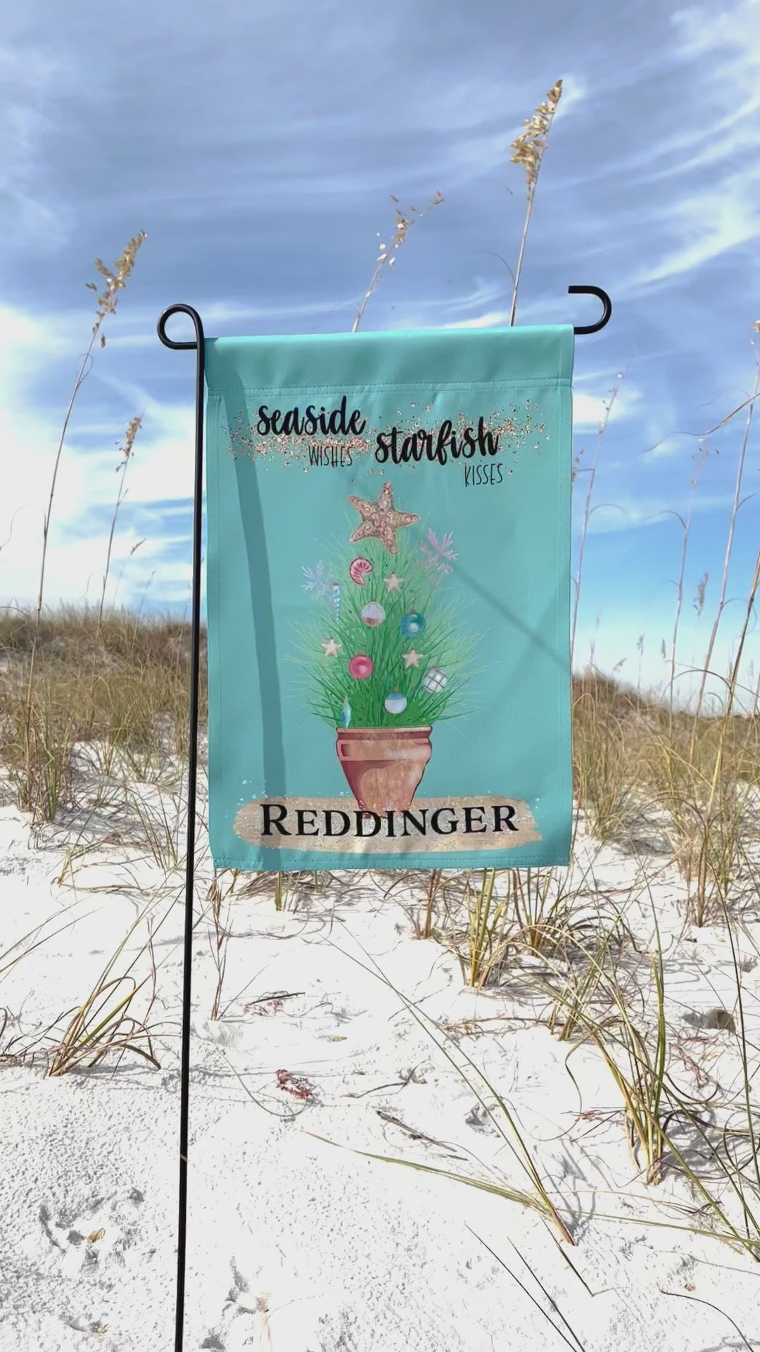 Personalized with a Last Name Flag. The blue green colored garden flag featuring potted ornamental grass adorned with Christmas tree bulb and topped with a golden starfish. The Flag reads 'Seaside Wishes Starfish Kisses'. Below the potted plant there is a large golden brushstroke with black bold text that reads 'Reddinger'. The flag is on a flag pole. A sand dune is visible in the background of the video. 