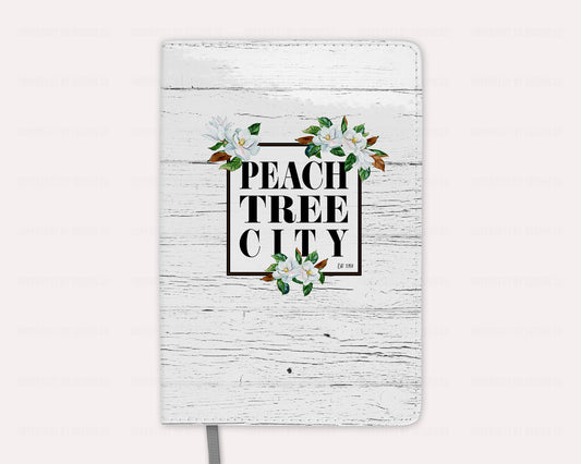 Elevate your desk decor with our Peachtree City magnolia notebook, featuring stunning magnolia blooms on a crisp white faux wood background, and Peachtree City name and date established. Perfect for organizing your notes and thoughts in style. | imperfect by design co