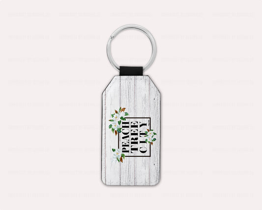 Peachtree City Magnolia Keychain - A stylish keychain with magnolia blooms on a white faux wood background, featuring the Peachtree City name, perfect for showing off your Peachtree City Pride on-the-go | imperfect by design co