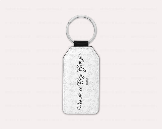 A faux leather keychain with a metal keyring featuring a grey whimsical leaves motif background and Peachtree City Georgia est. 1959 in the foreground. | imperfect by design co